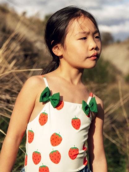 Young girl wearing a white top with strawberry patterns and green bows outdoors.