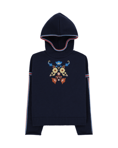 Two women sitting in a snowy landscape, wearing embroidered clothing, a true tribute to Norwegian heritage. This hooded sweater is crafted from 100% Norwegian wool, sourced from free-ranging sheep, and features embroidered motifs inspired by the traditional Norwegian Bunad of Telemark, Hallingdal, and Gudbrandsdalen.