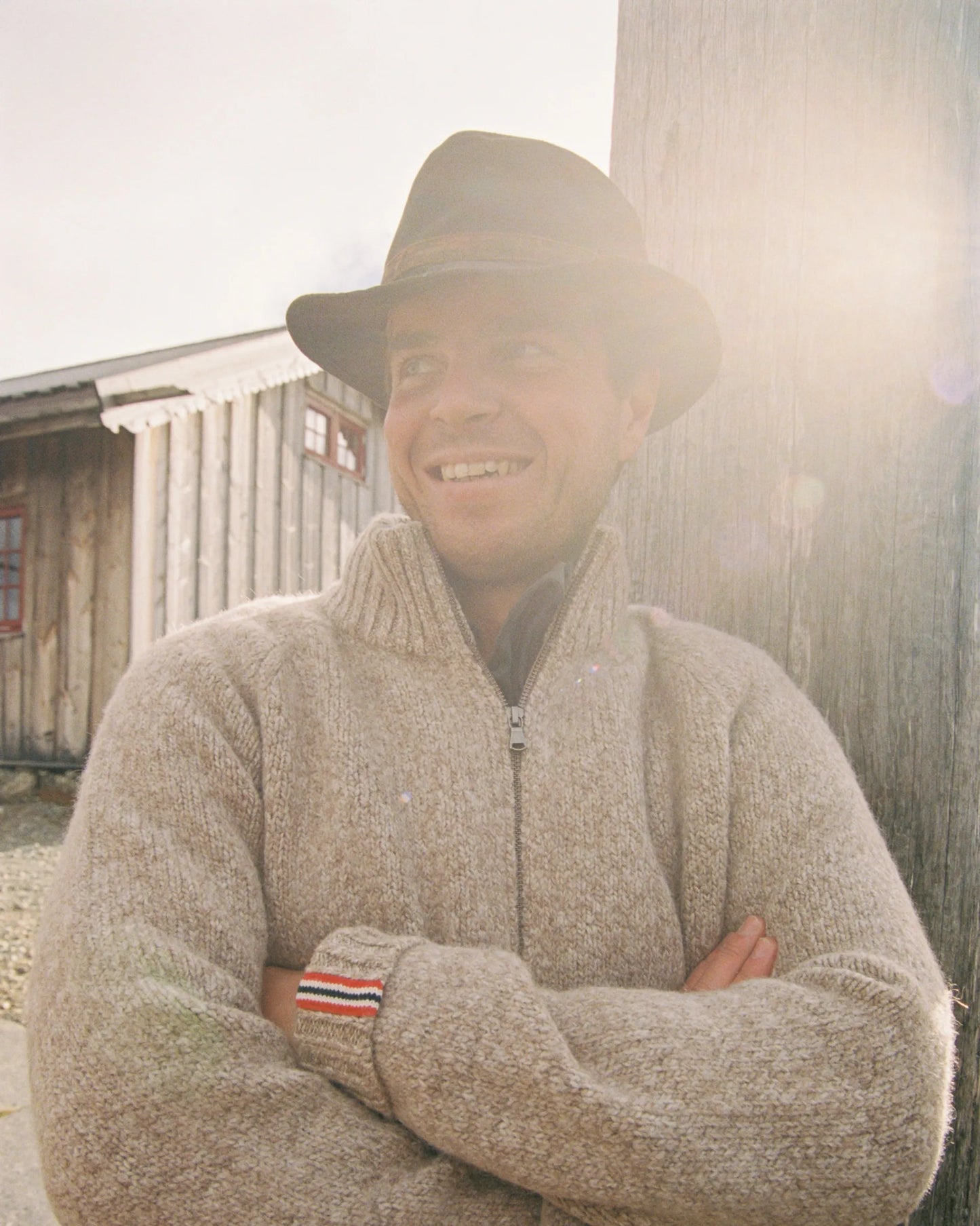 Man wearing a beige sweater with a Norwegian flag detail on the wrist, standing against a wooden building. Scandinavian Men's fashion found in Los Angeles at Huset Shop your home for all things Scandinavian and men's outdoor gear shopping in Venice California.