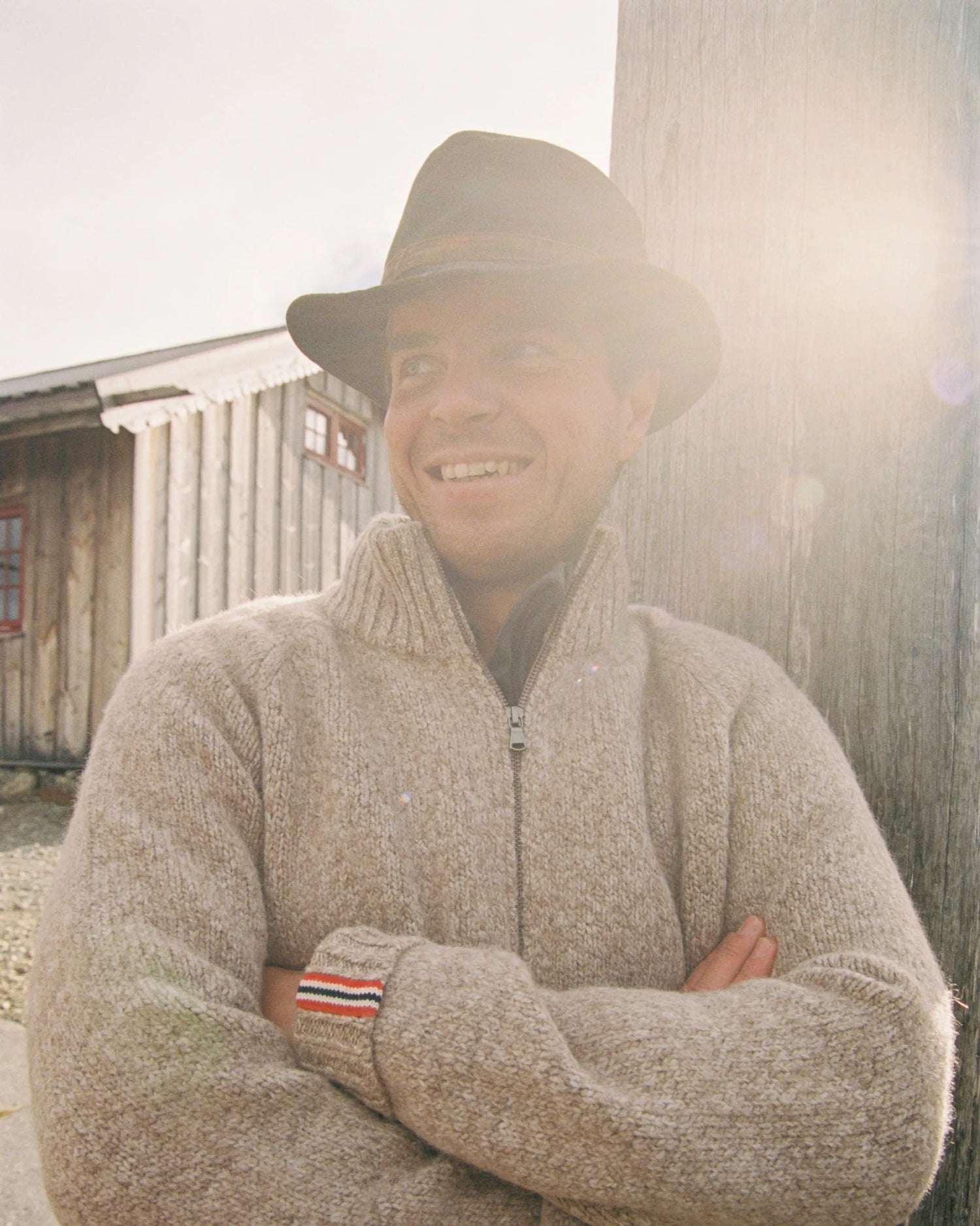 Man wearing a beige sweater with a Norwegian flag detail on the wrist, standing against a wooden building. Scandinavian Men's fashion found in Los Angeles at Huset Shop your home for all things Scandinavian and men's outdoor gear shopping in Venice California.