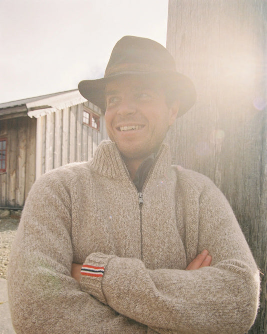 Man wearing a beige sweater with a Norwegian flag detail on the wrist, standing against a wooden building. Scandinavian Men's fashion found in Los Angeles at Huset Shop your home for all things Scandinavian and men's outdoor gear shopping in Venice California.