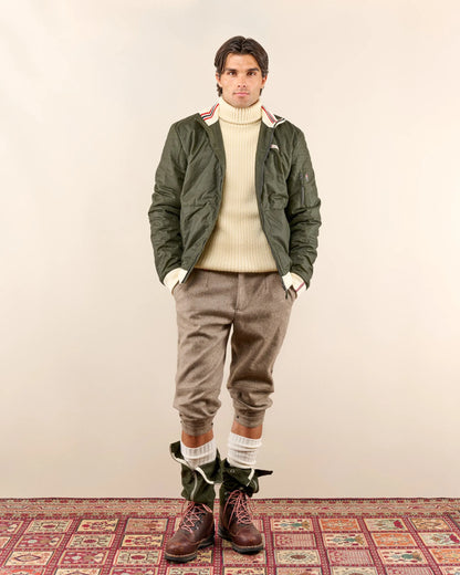 Man wearing a green jacket, beige sweater, brown pants, and boots on a patterned rug. Norwegian camping or hiking, or anything involving the outdoors you will love this jacket. Lightweight durable polyamide face, 120 grams Norwegian Woolinside insulation, Cuffs, waist and neck knitted in pure merino wool, Antarctic Tartan lining
Amundsen Peak patches on upper sleeves. He is using a camping stove to make some coffee surrounded by trees.