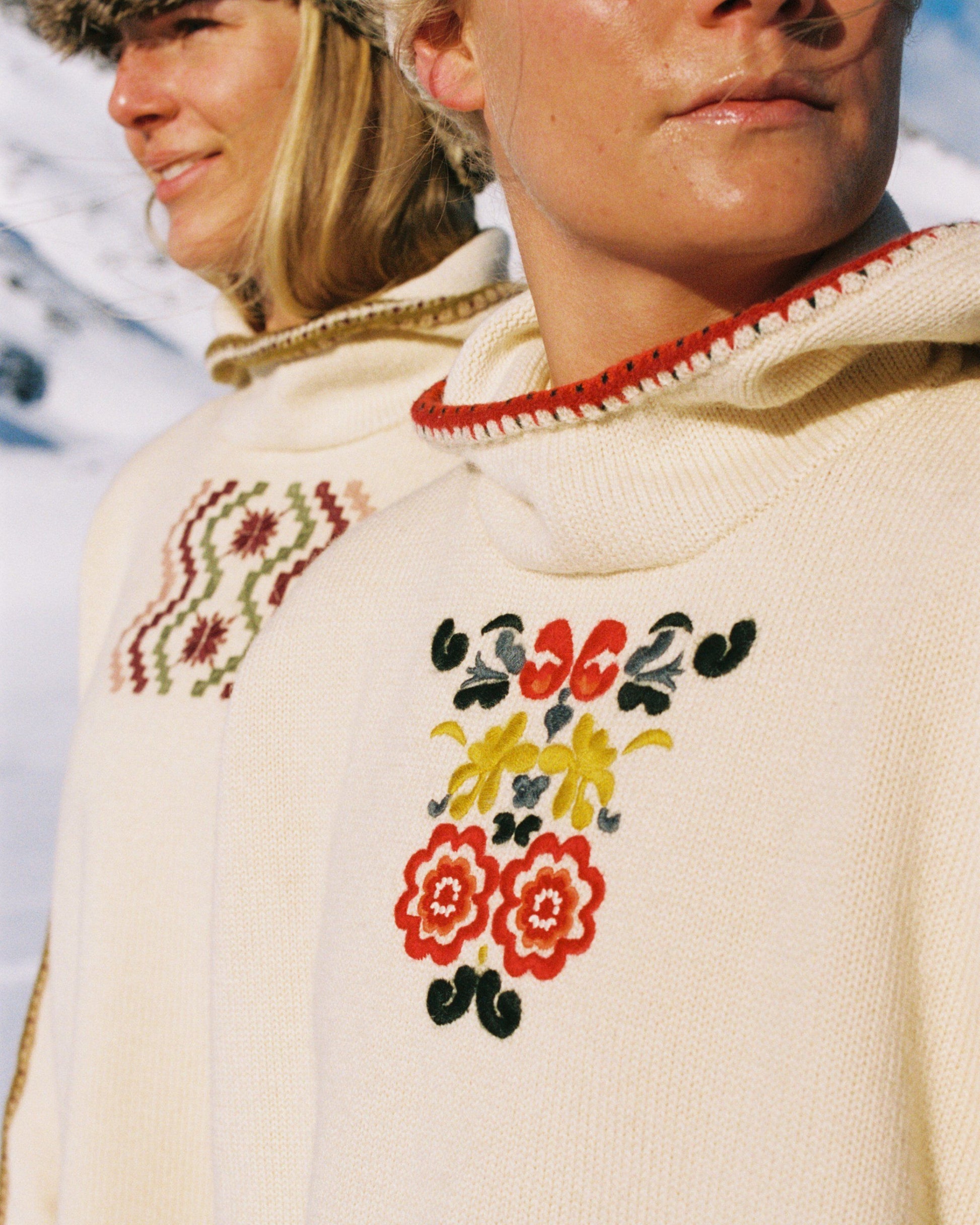 Two women sitting in a snowy landscape, wearing embroidered clothing, a true tribute to Norwegian heritage. This hooded sweater is crafted from 100% Norwegian wool, sourced from free-ranging sheep, and features embroidered motifs inspired by the traditional Norwegian Bunad of Telemark, Hallingdal, and Gudbrandsdalen.