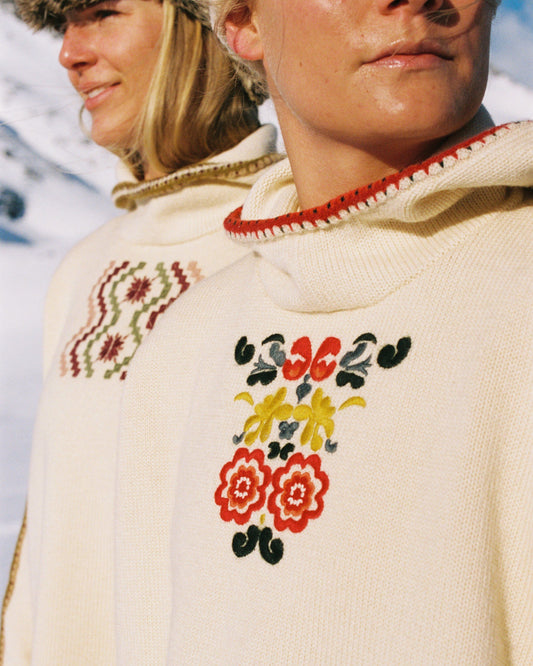Two women sitting in a snowy landscape, wearing embroidered clothing, a true tribute to Norwegian heritage. This hooded sweater is crafted from 100% Norwegian wool, sourced from free-ranging sheep, and features embroidered motifs inspired by the traditional Norwegian Bunad of Telemark, Hallingdal, and Gudbrandsdalen.