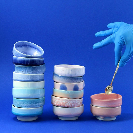Stacked ceramic dip dish bowls with a hand in a blue glove holding a spoon over one of them against a blue background. Studio Arhoj